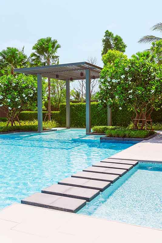 A serene outdoor swimming pool with stepping stones leading across the water. Lush greenery and well-manicured bushes surround the pool. A wooden pergola stands by the poolside, providing shade. Palm trees are visible in the background under a clear sky after edit.