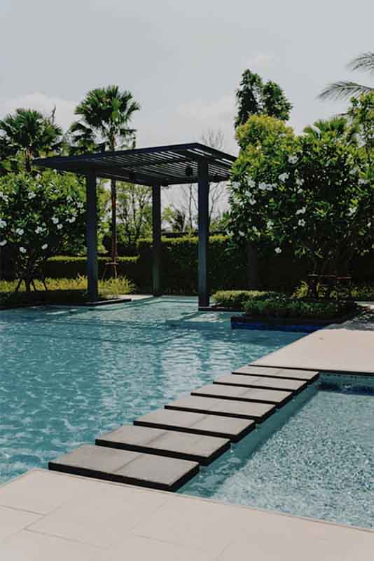 A serene outdoor swimming pool with stepping stones leading across the water. Lush greenery and well-manicured bushes surround the pool. A wooden pergola stands by the poolside, providing shade. Palm trees are visible in the background under a clear sky before edit.