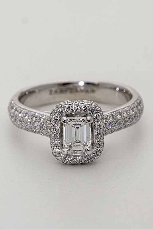 A close-up image of a diamond ring with a rectangular-cut center diamond, surrounded by a halo of smaller diamonds. The band is also encrusted with small diamonds, giving it a sparkling appearance. Before edit.