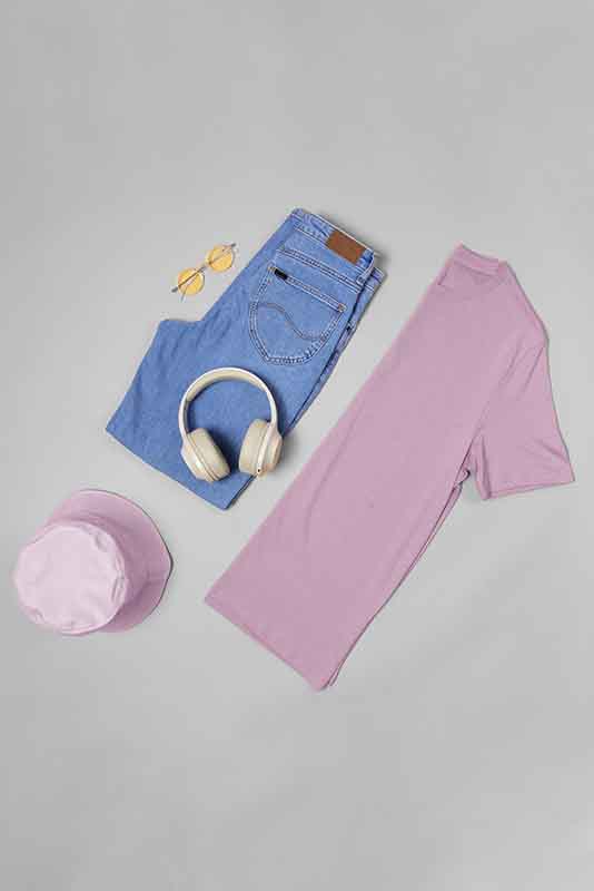 Flat lay of casual outfit: folded blue jeans with brown leather patch, light purple t-shirt, light purple bucket hat, beige over-ear headphones, and yellow-tinted round sunglasses, before edit.