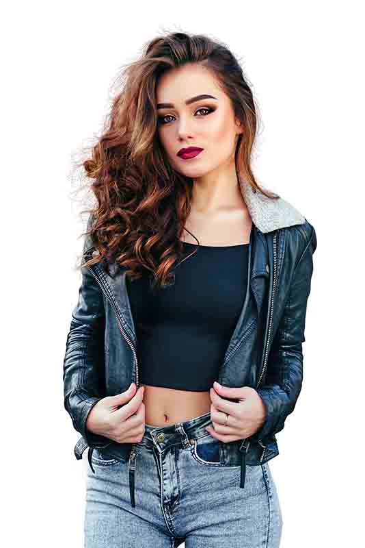 A woman with long, wavy brown hair stands confidently, wearing a black crop top, a black leather jacket with a shearling collar, and blue jeans. She has bold makeup and is holding the edges of her jacket with both hands. The background is transparent, perfect for showcasing our photo editing service.