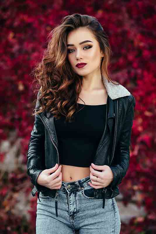 A woman with wavy brown hair and makeup poses confidently in front of a vibrant red foliage background. She wears a black crop top, blue jeans, and a black leather jacket with a shearling collar, slightly open with her hands holding the jacket.