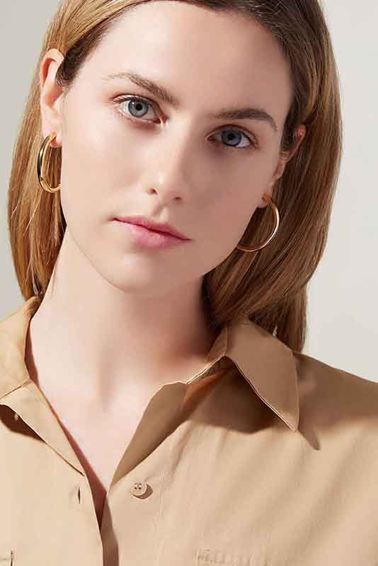 A women with light skin and straight blonde hair is facing the camera after edit. The individual is wearing gold hoop earrings and a beige collared shirt. The background is light and plain. Their expression is neutral.