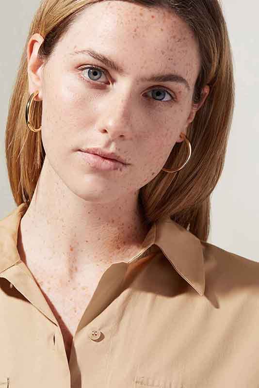 A women with light skin and straight blonde hair is facing the camera before edit. The individual is wearing gold hoop earrings and a beige collared shirt. The background is light and plain. Their expression is neutral.