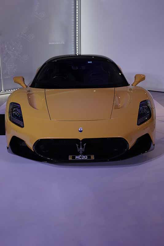 A yellow Maserati MC20 sports car before edit is showcased in a well-lit showroom, with its sleek and aerodynamic design prominently displayed. The car's front view highlights its bold grille and iconic trident logo.