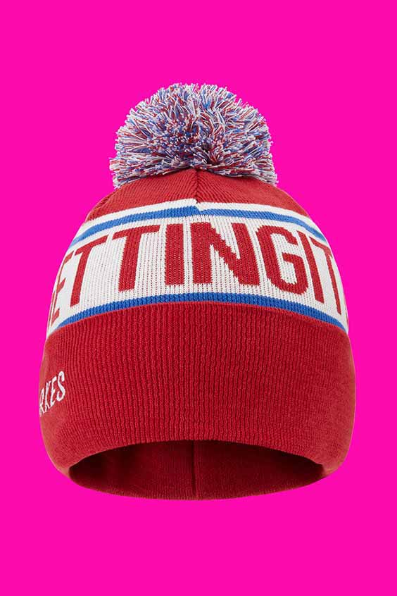 A red beanie hat with a colorful pom-pom on top. The hat features a blue and white band with text in red lettering on the front. The background is a bright pink shade.