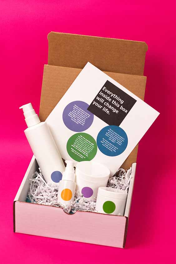 A cardboard box filled with skincare products, including bottles and jars, each labeled with a colored dot. An accompanying card reads, "Everything inside this box will change your life," along with other text and colored circles. The background is vividly pink.